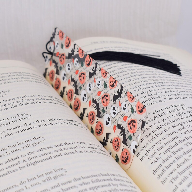 May include: A clear plastic bookmark with a Halloween theme. The bookmark features a repeating pattern of orange pumpkins, black bats, and white skulls. The bookmark has a black tassel.