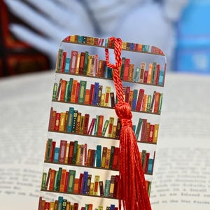 Peut inclure: Un marque-page à pompon rouge avec un design en plastique transparent représentant une bibliothèque remplie de livres colorés. Ce marque-page est parfait pour les bibliophiles et ajoute une touche de fantaisie à toute expérience de lecture.