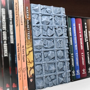 May include: A gray 3D printed bookend shaped like a cage filled with dice. The bookend is sitting on a bookshelf with several books visible. The books are titled "Monster Manual II", "Maera Gazetteer", "Hoard of the Dragon Queen", and "Guildmaster's Guide to Ravnica".