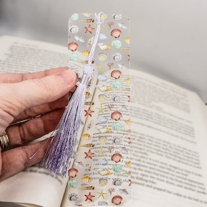 May include: A clear bookmark with a purple tassel. The bookmark is decorated with a pattern of seashells, starfish, and coral in shades of pink, orange, yellow, and blue.
