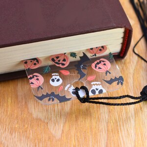 May include: A clear plastic bookmark with a Halloween theme. The bookmark features a repeating pattern of orange pumpkins, black bats, and white skulls. The bookmark has a hole at the top for a string or ribbon.