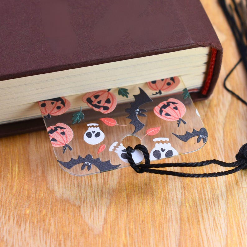 May include: A clear plastic bookmark with a Halloween theme. The bookmark features a repeating pattern of orange pumpkins, black bats, and white skulls. The bookmark has a hole at the top for a string or ribbon.