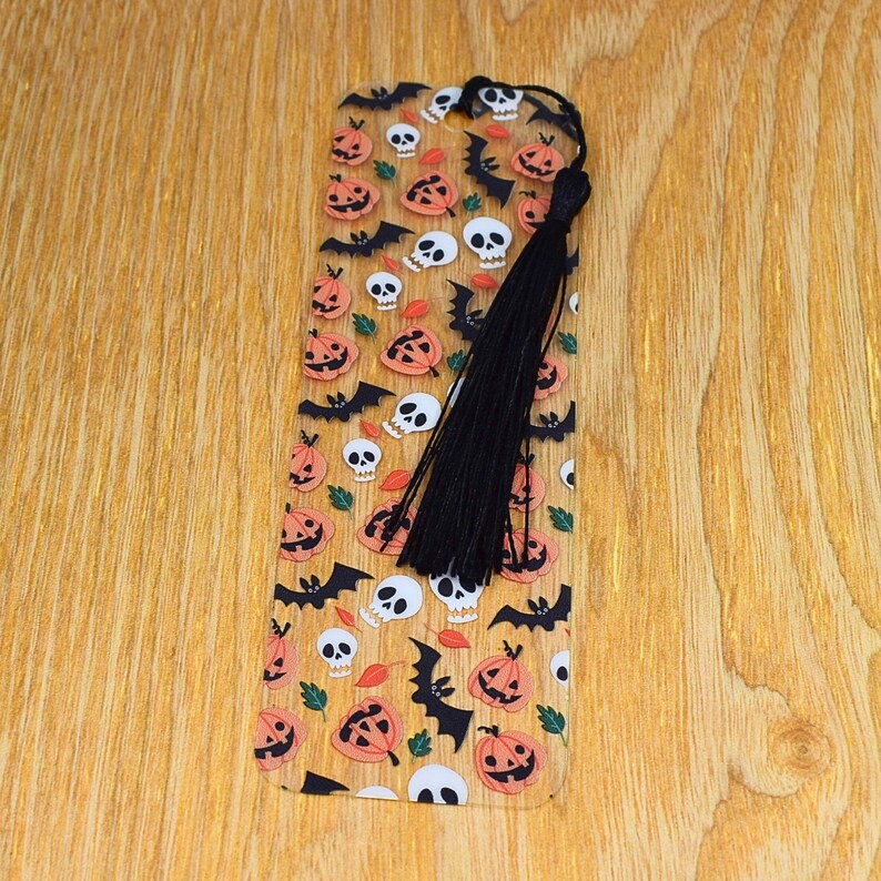 May include: A clear plastic bookmark with a Halloween theme. The bookmark features a repeating pattern of orange pumpkins, black bats, and white skulls. It has a black tassel.