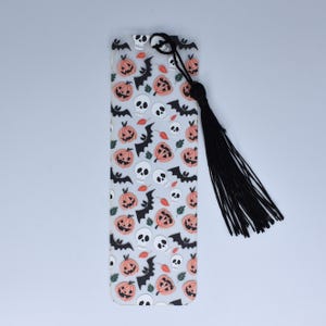 May include: A clear plastic bookmark with a black tassel. The bookmark features a repeating pattern of orange pumpkins, white skulls, and black bats.