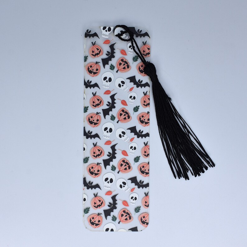 May include: A clear plastic bookmark with a black tassel. The bookmark features a repeating pattern of orange pumpkins, white skulls, and black bats.