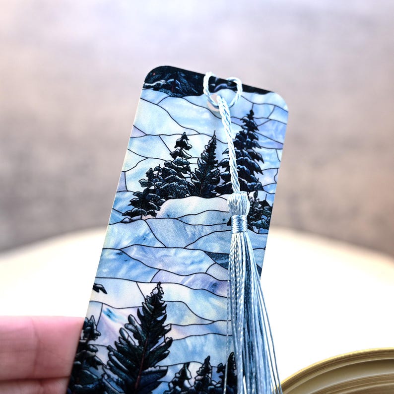 May include: A blue and white stained glass bookmark with a winter scene featuring trees and snow. The bookmark has a light blue tassel.