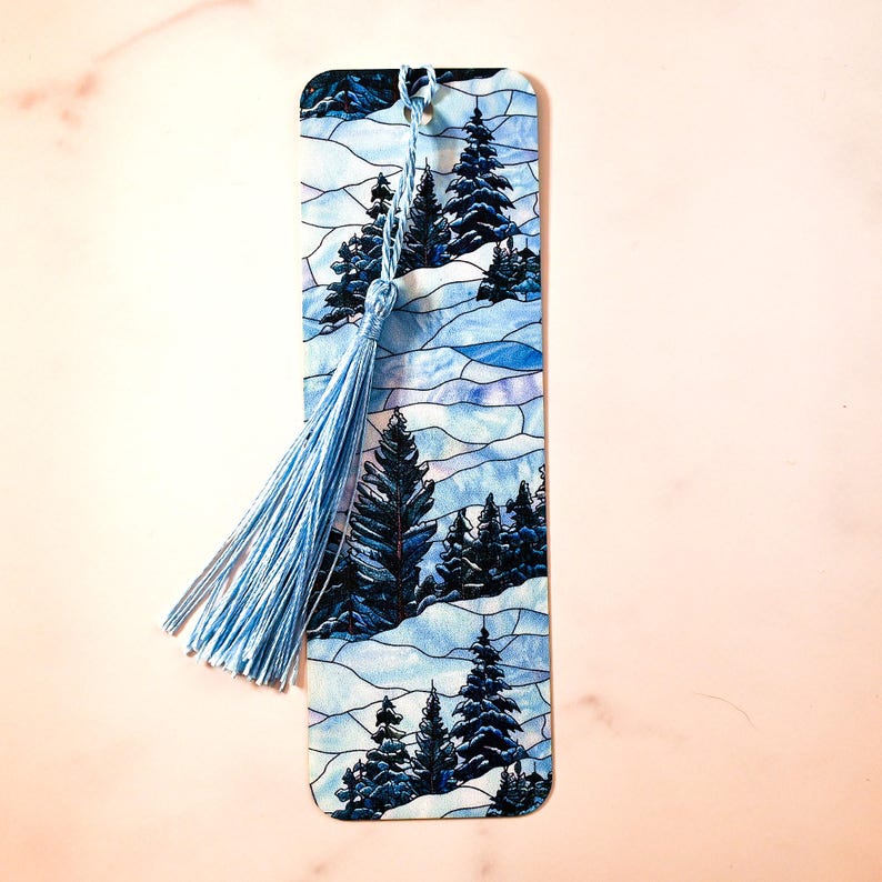 May include: A blue and white bookmark with a stained glass design of a winter forest scene. The bookmark has a blue tassel.