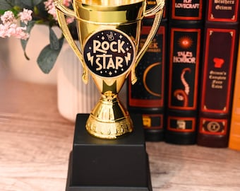 May include: A gold-colored trophy with a black base. The trophy has a circular emblem with the words "ROCK STAR" in white lettering. The trophy is set against a backdrop of books and a vase of flowers.