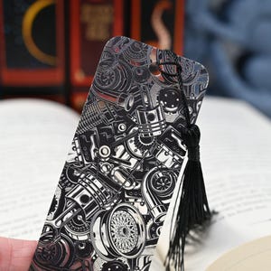 May include: A black and silver bookmark featuring an intricate design of engine parts, including pistons, gears, and wheels. The bookmark has a black tassel attached at the top. The background shows a book and other books.