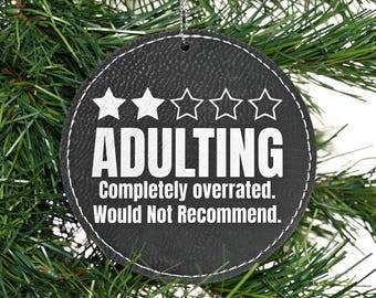 May include: A black leather-like Christmas ornament with a white star rating of one out of five stars and the text "Adulting Completely overrated. Would Not Recommend."