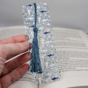May include: A clear bookmark with a blue tassel. The bookmark features a repeating pattern of white sharks, blue coral, and blue stars on a white background.