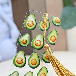 May include: A clear acrylic bookmark featuring a pattern of smiling avocado halves. Each avocado has a green exterior and a smiling orange pit. A gold tassel hangs from the top of the bookmark.