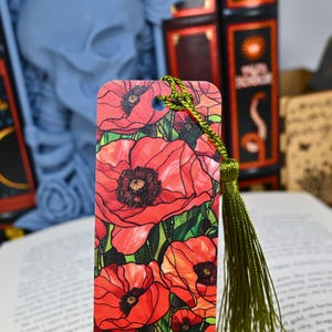 May include: A rectangular bookmark featuring a vibrant floral design of red poppies with green leaves. The bookmark has a green tassel attached to the top. The background includes open books and decorative items.