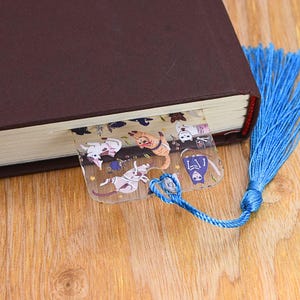 May include: A clear plastic bookmark with a blue tassel. The bookmark features a colorful illustration of cats and a skeleton wearing a blue robe.