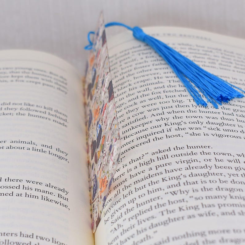 May include: A colorful bookmark with a blue tassel. The bookmark is made of paper and has a collage of images and text. The text on the bookmark reads "Because our King's only daughter is sick unto death."