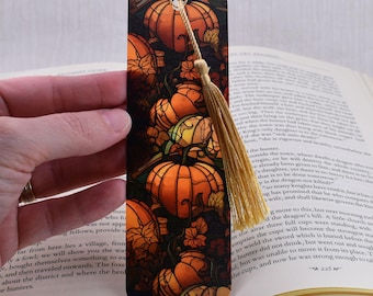 Puede incluir: Un marcapáginas decorativo con un diseño de calabaza. El marcapáginas presenta un patrón repetido de calabazas naranjas con contornos negros y hojas verdes. Una borla dorada cuelga de la parte superior. El marcapáginas está sobre un libro abierto.