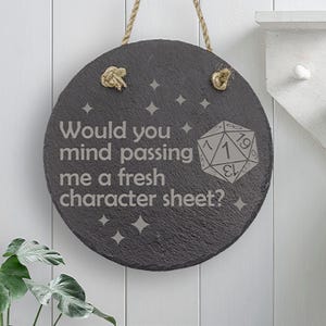 May include: Circular dark grey slate sign with the text "Would you mind passing me a fresh character sheet?" and a die graphic. The sign is hung by a natural rope. Decorative stars are also etched on the sign.
