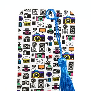 May include: A white bookmark with a blue tassel. The bookmark is covered in a pattern of colorful cameras and other photography-related icons.