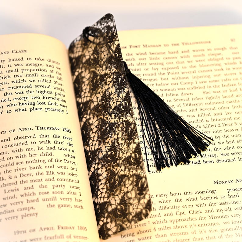 May include: A black lace bookmark with a black tassel. The bookmark is laying on a page of a book with text about the Lewis and Clark expedition.