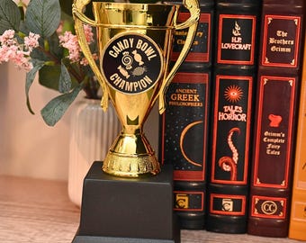 May include: A gold-colored trophy with a black base. The trophy has a cup-shaped top with handles and the words "CANDY BOWL CHAMPION" on a black circular emblem. The trophy is displayed next to a row of books with titles like "Tales of Horror" and "The Brothers Grimm".