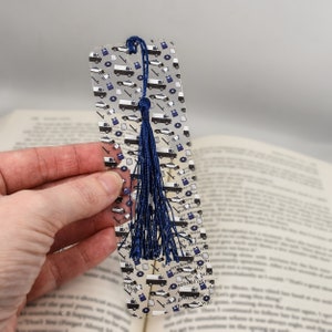 May include: A clear plastic bookmark with a blue tassel. The bookmark is decorated with a repeating pattern of police cars, badges, and other police-related imagery.