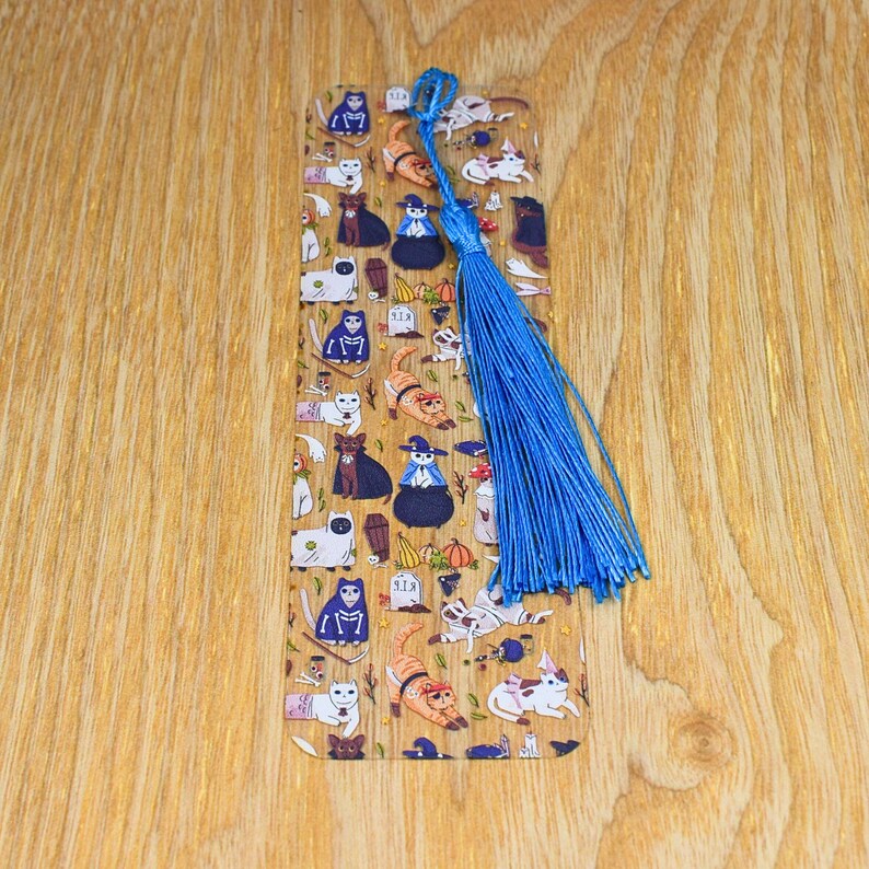 May include: A clear acrylic bookmark with a Halloween cat design. The bookmark features a variety of cats dressed in costumes, including witches, skeletons, and pumpkins. The bookmark has a blue tassel.