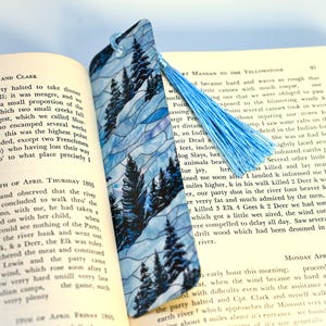 May include: A blue and white stained glass bookmark featuring a wintery forest scene with tall pine trees. The bookmark has a blue tassel attached to the top.