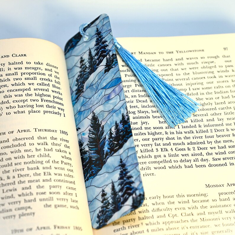 May include: A blue and white stained glass bookmark featuring a wintery forest scene with tall pine trees. The bookmark has a blue tassel attached to the top.