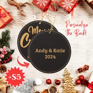 Może przedstawiać: Dwie czarne sk&oacute;rzane ozdoby choinkowe ze złotym przeszyciem. Ozdoba z przodu ma napis "Merry Christmas" złotymi literami. Ozdoba z tyłu ma napis "Andy & Katie 2024" złotymi literami.