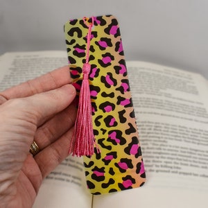 May include: A rectangular bookmark with a leopard print design in yellow, pink, and black. The bookmark has a pink tassel attached at the top. The bookmark is held over an open book.