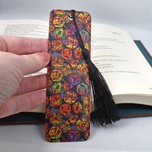 Puede incluir: Un marcapáginas negro con un diseño de dados de colores iridiscentes. El marcapáginas tiene un borla negra.