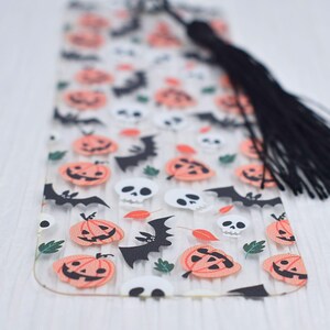 May include: A clear plastic bookmark with a Halloween theme. The bookmark features a repeating pattern of orange pumpkins, black bats, and white skulls. The bookmark has a black tassel.