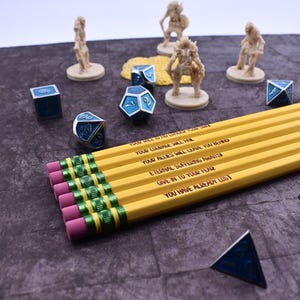 May include: Five yellow pencils with black and green stripes and pink erasers. The pencils have motivational messages printed on them: "Your will is no longer your own", "Your courage will fail", "Your allies will leave you behind", "Eternal suffering awaits", "Give in to your fear", "You have already lost".