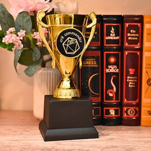May include: A gold-colored trophy with a black base. The trophy has a cup-shaped top with handles and a circular emblem that reads "crit commander" and "20". Books with titles like "Tales of Horror" and "The Brothers Grimm" are in the background.