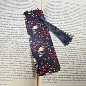 May include: A black bookmark with a dark background featuring skulls and red berries. A navy blue tassel hangs from the top. The bookmark is placed on top of an open book.