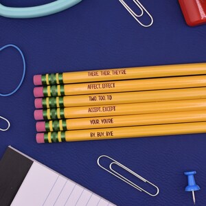 May include: Six yellow pencils with pink erasers and green metal ferrules. Each pencil features black printed text: THERE. THEIR. THEY'RE, AFFECT. EFFECT, TWO. TOO. TO, ACCEPT. EXCEPT, YOUR. YOU'RE, and BY. BUY. BYE, arranged in a parallel horizontal stack.