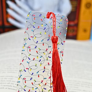May include: A decorative bookmark with a festive design. The bookmark features a pattern of colorful Christmas lights on a silver background. A long red tassel hangs from the top. The bookmark is designed for use in books.