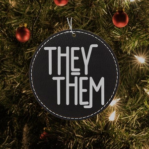 May include: Black leather Christmas bauble with white stitching around the edge. The bauble has the words "THEY THEM" written in white on a black background.
