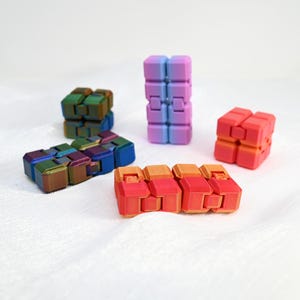 May include: Five colourful fidget toys made of plastic. The toys are in different shapes and sizes, including a cube, a rectangle, and a long, thin shape. The colours of the toys are pink, red, orange, green, and blue.
