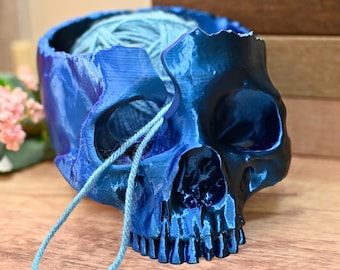 May include: A blue skull-shaped yarn bowl with a ball of blue yarn inside. The yarn is threaded through a hole in the skull and hangs down.