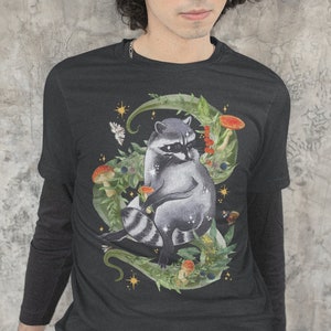 May include: Black long-sleeve t-shirt with a graphic print of a raccoon sitting in a circle of leaves and mushrooms. The raccoon has a black mask and white stripes on its tail. The design is surrounded by stars and other foliage.