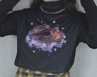 May include: A black long-sleeved shirt with a graphic of a fawn lying in a bed of purple flowers. The shirt is paired with a yellow and black plaid skirt.