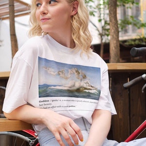 May include: A woman wearing a white t-shirt with a painting of a goddess on the front. The shirt has the word "Goddess" and a definition printed on the bottom. She is sitting on a red bicycle with a blue and white striped skirt.
