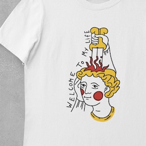 May include: White t-shirt with a graphic of a person with a knife through their head. The text "WELCOME TO MY LIFE" is written vertically on the left side of the graphic.
