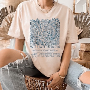 May include: A beige t-shirt with a blue floral print and the text "WILLIAM MORRIS COTTON PRINTS EXHIBITION VICTORIA & ALBERT MUSEUM FEB. 10TH TO APR. 18TH ADMISSION FREE NEAEST UNDERGROUND STATION SOUTH KENSINGTON"