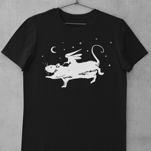 Puede incluir: Camiseta negra con un gr&aacute;fico blanco de una hada montando una rata bajo una luna creciente y estrellas.
