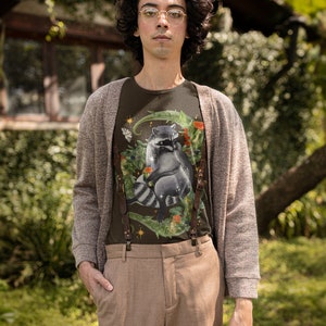 May include: A person wearing a brown cardigan over a dark green t-shirt with a raccoon graphic. The t-shirt has a floral design and a star. The person is wearing brown pants and suspenders.