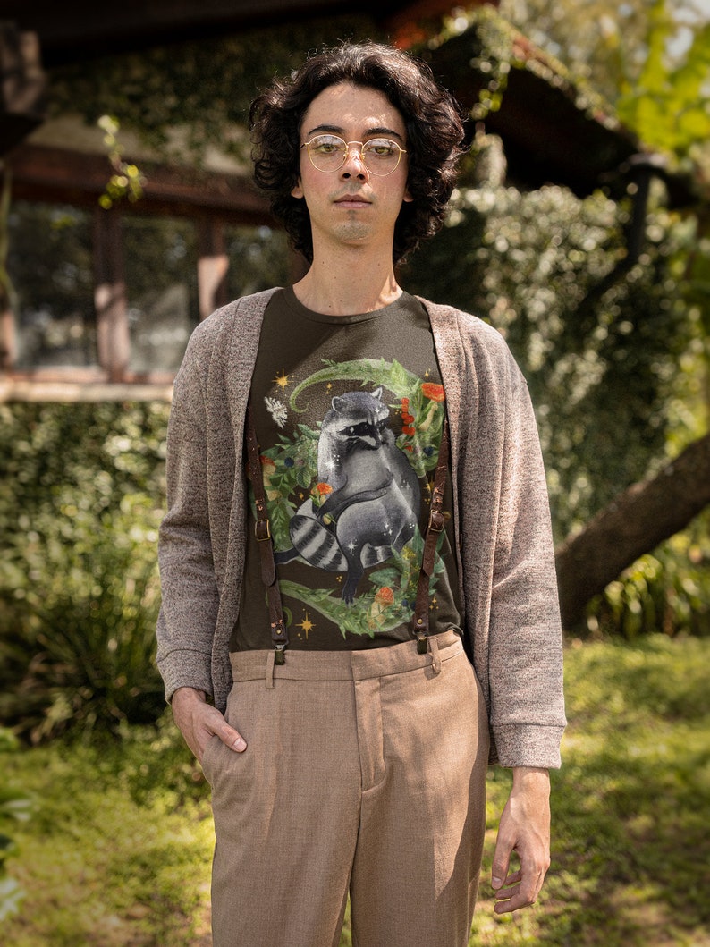 May include: A person wearing a brown cardigan over a dark green t-shirt with a raccoon graphic. The t-shirt has a floral design and a star. The person is wearing brown pants and suspenders.