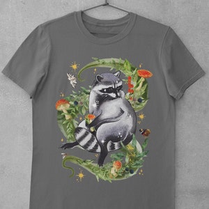 May include: A dark gray t-shirt with a graphic of a raccoon sitting in a circle of green leaves, mushrooms, and butterflies. The raccoon is gray and black with a white mask.