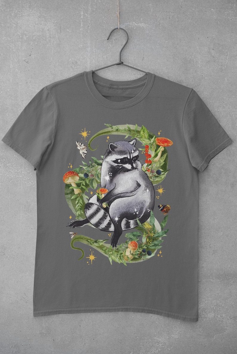 May include: A dark gray t-shirt with a graphic of a raccoon sitting in a circle of green leaves, mushrooms, and butterflies. The raccoon is gray and black with a white mask.